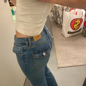 Blå bootcut jeans från Gina Tricot - Snygga blå jeans från Gina Tricot med bootcut-modell och låg midja. Jeansen har klassiska fickor fram och bak, varav bakfickorna har lock och knappdetalj. Tillverkade i mjukt denimtyg med ljus tvätt och kontrastsömmar.