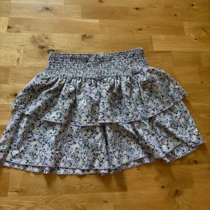 Ljuslila blommig volangkjol - Supersöt kort kjol med blommigt mönster i ljuslila, vitt och grönt. Kjolen har bred smock i midjan och dubbla volanger som ger en flowy känsla. Perfekt för varma dagar och enkel att styla med en enkel topp.