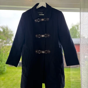 Svart rock från Sportmax Code - Stilren svart rock från Sportmax Code med snygga spännen framtill och klassisk krage. Jackan har två fickor och är helfodrad, perfekt för kyliga dagar. Långärmad och rak modell som ger en clean och tidlös look.