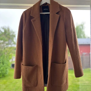 Brun kappa från Monki, storlek S - Säljer en snygg brun kappa från Monki i storlek S. Jackan har klassisk krage, två stora fickor framtill och är helfodrad. Perfekt för dig som gillar stilrena och tidlösa plagg. Materialet är mjukt och känns lyxigt.
