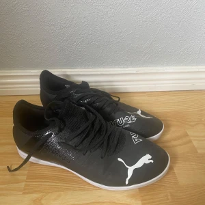 Svarta Puma Future fotbollsskor - Svarta Puma Future fotbollsskor med vit logga och text på ovansidan. Skorna har snörning och en strumpliknande krage för extra stöd. Snygg och sportig design med platt sula, perfekta för inomhusspel. Tillverkade i syntetmaterial.
