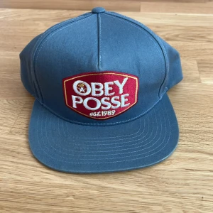 Blå Obey keps med röd patch - Snygg blå keps från Obey med stor röd och vit broderad patch framtill där det står 'OBEY POSSE est. 1989'. Kepsen har klassisk snapback-stängning och är tillverkad i 100% bomull. Perfekt för dig som gillar streetwear och vill sticka ut.