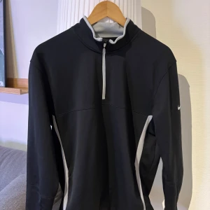 Nike Golf svart tröja half zip M - Svart långärmad Nike Golf Therma-Fit tröja med grå detaljer och halv dragkedja framtill. Snygg sportig look med vit Nike-logga på ärmen. Perfekt för golf eller träning, standard fit och tillverkad i funktionsmaterial.  
