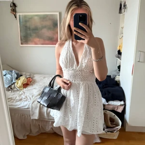 Vit kort halterneckklänning med broderi - Supersöt vit kort klänning med halterneck och öppen rygg. Klänningen har broderade detaljer och smock i midjan för en snygg passform. 