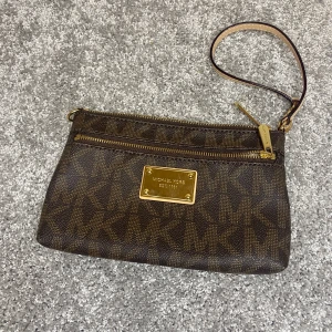 Brun Michael Kors kuvertväska - Snygg brun kuvertväska från Michael Kors med guldiga detaljer och logomönster över hela väskan. Väskan har en avtagbar handledsrem och dragkedja upptill samt en extra ficka med dragkedja framtill. Perfekt för att bära det viktigaste med stil.