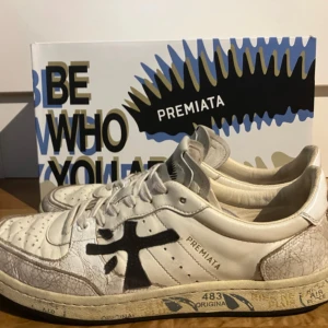 Premiata vita sneakers med svart logga - Snygga vita sneakers från Premiata med svart logga på sidan och coola detaljer på sulan. Skorna har en unik krackelerad effekt på lädret, perforerad tå och klassisk snörning. Storlek 43! Skorna är ok skick