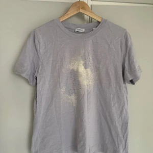 Ljusgrå t-shirt med guldtryck Esprit - Snygg ljusgrå t-shirt från Esprit med korta ärmar. Framsidan har ett abstrakt guldtryck och små silvriga nitar som ger en cool touch. Tillverkad i mjuk bomull och har en klassisk passform. Perfekt för dig som gillar stilrena men ändå unika plagg.