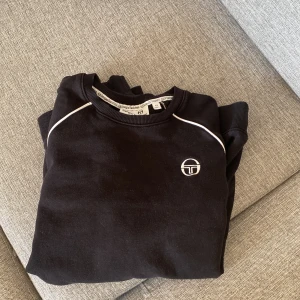 Svart sweatshirt från Sergio Tacchini - Svart sweatshirt från Sergio Tacchini med vita detaljer längs ärmarna och axlarna samt logga på bröstet. Tröjan har rund halsringning och långa ärmar med ribbade muddar. Perfekt för en sportig och avslappnad stil.