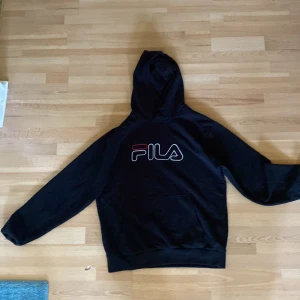 Svart Fila hoodie med logga - Svart hoodie från Fila med stor, vit och röd logga på bröstet. Klassisk känguruficka framtill och huva. Mjuk och skön bomullsblandning, perfekt för chill eller streetstyle. Långärmad och normal passform. Kan till komma katthår för finns katt i huset