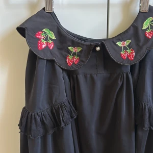 Svart topp med jordgubbsbroderi - Supersöt svart topp med stor krage dekorerad med broderade jordgubbar och en pärlknapp. Vida långärmar med volangdetaljer och spetskant. Toppen har en loose passform och känns både lekfull och unik.