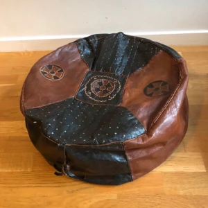 Stor oanvänd läderpuff med fint mönster från Västafrika - Unik rund sittpuff i brunt och svart läder med broderade detaljer och mönster på ovansidan. Säljes ostoppad och har dragkedja på undersidan. Perfekt för dig som gillar boho eller vintage vibes. 55cm i diameter och 20cm hög när den är stoppad (skumgummi eller tidningspapper).
