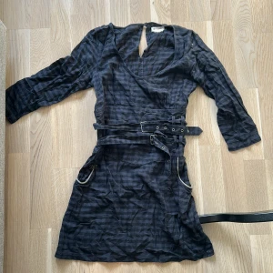 Mörkgrå rutig kortklänning med bälte - Snygg mörkgrå rutig klänning från wrangler med långa ärmar och markerad midja med dubbla bälten och metallspännen. Klänningen har djup rundringning och coola detaljer vid fickorna. Köpte från selpy men var alldeles för liten för mig. Men den är jätte cool, kan ta fler bilder om efterfrågas