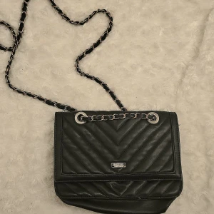 Svart quiltad axelväska från Don Donna - Snygg svart axelväska från Don Donna i quiltat skinn med kedjerem och silverdetaljer. Väskan har magnetlås, dragkedjeficka på baksidan och klassisk logga framtill. Perfekt storlek för det viktigaste och ger en stilren vibe till din outfit.