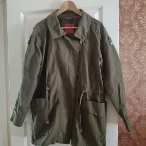 Olivgrön parkajacka från edc, L - Snygg olivgrön parkajacka från edc med dragkedja, stora fickor och justerbar midja med snörning. Jackan har klassisk krage och långa ärmar med knappdetaljer. Perfekt för lager-på-lager och chill vår- eller höstdagar.