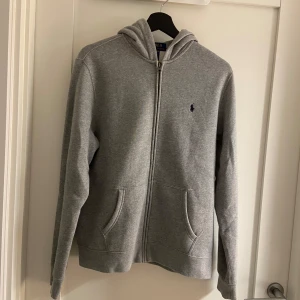 Grå hoodie från Polo Ralph Lauren  - Klassisk grå hoodie från Polo Ralph Lauren. Tröjan har dragkedja, huva och två fickor framtill. Liten mörkblå logga broderad på bröstet. Storlek 18-20 motsvarar ungefär S i vanlig storlek 