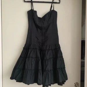Svart volangklänning med smala axelband - Supersnygg svart kortklänning med smala axelband och volanger i flera lager. Klänningen har en markerad byst och en utställd kjol som ger en cool och lekfull look.