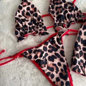 Leopardmönstrad bikini med kjol - Trendigt bikini-set med leopardmönster i svart och bruna toner, röda kanter och smala knytband. Setet består av trekantsbikini, trosa med höga skärningar och matchande kjol. Perfekt för dig som vill sticka ut på stranden.