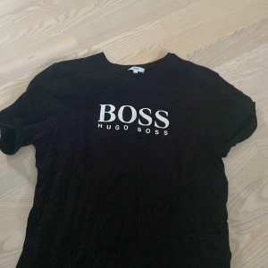 Svart t-shirt från Hugo Boss - Svart t-shirt från Hugo Boss med klassisk rund hals och vit logotyptryck på bröstet. Enkel och stilren design som passar perfekt till jeans eller shorts. Tillverkad i mjuk bomull för skön känsla hela dagen.