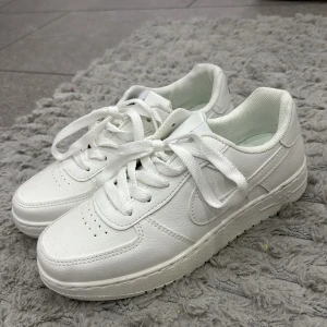Nike Air Force 1 vita sneakers - Klassiska Nike Air Force 1 sneakers i helvitt skinn med rund tå och platt sula. Skorna har tydlig Nike-logga bak och på plösen, samt perforerade detaljer framtill för extra ventilation. Perfekta för dig som gillar en clean och tidlös look.
