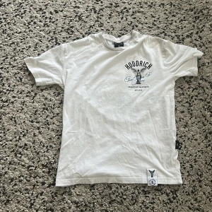  Hoodrich T-shirt  ängeltryck - Vit t-shirt från Hoodrich i bomull med svart och blått tryck framtill och stort ängelmotiv på ryggen. Texten 'From Nothing To Something' i blått och cool street vibe. Klassisk passform och rund hals. Perfekt för dig som gillar streetwear och snygga prints.