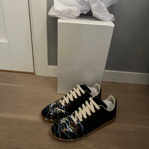 Maison Margiela sneakers med färgstänk - Svarta sneakers från Maison Margiela med unika färgstänk i blått, gult, vitt och rött. Skorna har vita snören, beige innersida och gummisula. Klassisk rund tå och låg profil, tillverkade i skinn med konstnärlig vibe. Skorna funkar på storlek 40,41