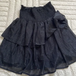 Svart volangkjol med glittriga detaljer - Supersnygg svart kortkjol med volanger och glittriga inslag. Kjolen har smock i midjan för en bekväm passform och ett skirt, lätt material som ger en cool look. Perfekt att styla med en crop top eller stickad tröja.