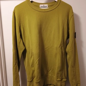 Olivgrön sweatshirt från Stone Island - Säljer en olivgrön sweatshirt från Stone Island med klassisk rund hals och ribbade muddar. Tröjan har den ikoniska Stone Island-loggan på vänster ärm. Tillverkad i mjuk bomull och har en clean, stilren look som passar perfekt till streetwear.