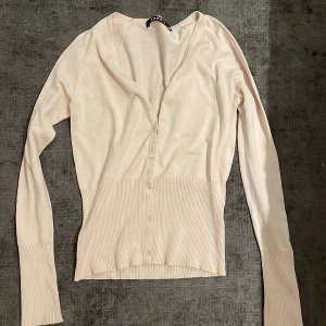 Ljusbeige kofta från Caroll - Supersöt ljusbeige kofta från Caroll med smickrande v-ringning och knappar framtill. Långärmad modell med bred ribbad mudd i midjan och vid ärmslut. Perfekt att slänga över en topp för en chill och stilren look.