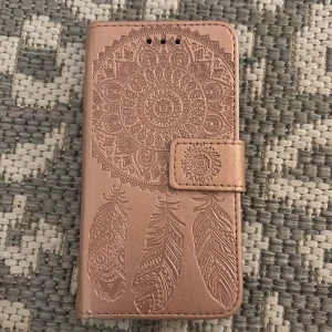 Rosa plånboksväska med mandalamönster - Snygg rosa plånboksväska till mobilen med detaljerat mandala- och fjädermönster. Väskan har flera kortfack och stängs med magnetknapp. Tillverkad i syntetmaterial med en mjuk, matt finish. Perfekt för dig som vill ha både stil och funktion i ett.