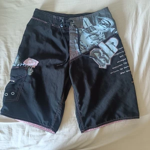 Svarta Rip Curl boardshorts med tryck - Coola svarta boardshorts från Rip Curl med stora tryck och textdetaljer i blått och vitt. Shortsen har snörning i midjan, praktiska fickor med öljett och kontrastsömmar i rosa. Perfekta för stranden eller skateparken.