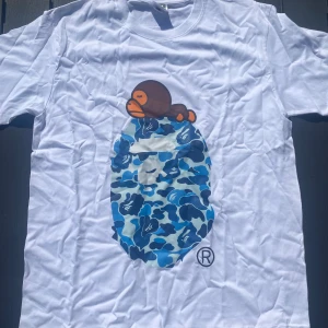 Vit BAPE t-shirt med blått camo-tryck - Large - Vit t-shirt med ett tryck av en anka ovanpå en blå kamouflagemönstrad form. Trycket har en distinkt och igenkännlig design. Knappt använd (2-3 ggr), tvättad efteråt. Storlek Large L
