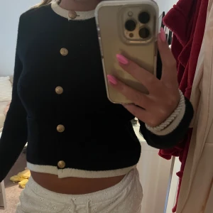 Svart kofta med guldknappar från Zara - Stilren svart kofta från Zara med kontrasterande vita kanter vid hals och nederkant. Koftan har snygga guldknappar framtill och två fickor. Långärmad och stickad i mjukt material, perfekt för lager-på-lager och en clean look.