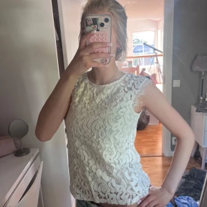 Vit spetsblus med ärmlös design - Supersöt vit blus i spets med ärmlös modell och rund halsringning. Blusen har fina spetsdetaljer över hela och en lätt transparent känsla. Perfekt att styla med jeans eller kjol för en fräsch look.