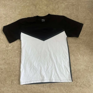 Nike svartvit t-shirt med V-design - Nike t-shirt i svart och vitt med snygg V-formad färgblock-design på framsidan. Klassisk rund hals och korta ärmar. Tillverkad i mjuk bomull, perfekt för en clean och sportig look. Diskret Nike-logga på bröstet.