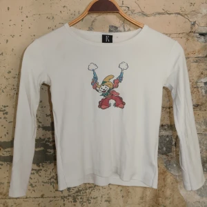 Vit långärmad tröja med clowntryck - Vit långärmad tröja från Réalisation Par med ett färgglatt tryck av en clown med två pistoler på bröstet. Tröjan har rund halsringning och är i mjukt bomullsmaterial, perfekt för dig som gillar unika och lekfulla prints.