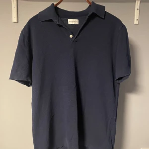 Marinblå pikétröja These Glory Days - Snygg marinblå pikétröja från These Glory Days i storlek L men passar jättebra för M. Klassisk krage, kort ärm och tre knappar framtill. Tröjan är gjord i bomull och har en stilren, enkel look som passar perfekt till jeans eller shorts.