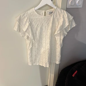 Vit blus från Holly & Whyte - Superfin vit blus från Holly & Whyte med snyggt strukturmönster och korta volangärmar. Blusen har rund halsringning och knäppning bak i nacken. Perfekt för dig som gillar en romantisk och stilren look. Lägger ut pga att den inte har kommit till användning💕