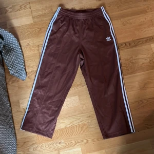 Bruna träningsbyxor från Adidas - Säljer ett par bruna träningsbyxor från Adidas med de klassiska tre vita ränderna längs sidorna. Byxorna har en elastisk midja och är tillverkade i ett syntetmaterial som ger en bekväm passform. Perfekta för en sportig look!