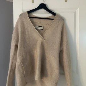 Beige stickad tröja By Malene Birger - Supermjuk och oversized beige stickad tröja från By Malene Birger. Tröjan har v-ringning och långa ärmar, perfekt för lager-på-lager och chill vibes. Den har en enkel och clean design som funkar till allt.