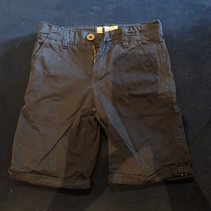 Svarta shorts från Hampton Republic - Klassiska svarta shorts från Hampton Republic med normal passform. De har fickor fram och bak, snygga bälteshällor och stängs med dragkedja och träfärgad knapp. Perfekta för sommardagar och enkla att matcha med det mesta.