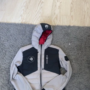 Grå cardiganjacka från Sail Racing XS - Säljer en grå och svart pufferjacka från Sail Racing i storlek XS. Jackan har huva med röd insida, dragkedja framtill och coola logodetaljer på bröstet och ärmen. Perfekt för kalla dagar och riktigt snygg streetstil.