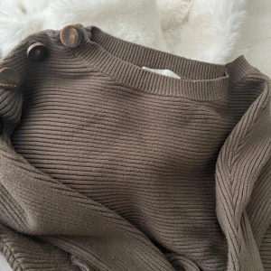 Brun ribbad stickad tröja med knappar - Säljer en brun-grön ribbad stickad tröja med stora dekorativa knappar längs axeln. Tröjan har en rund halsringning och är i ett mjukt, skönt material som passar perfekt för kyligare dagar. Enkel att styla med jeans eller kjol.