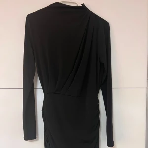 Svart långärmad klänning  - Snygg svart kortklänning med långa ärmar och draperad detalj över axeln och sidan. Klänningen har en figurnära passform och är tillverkad i ett mjukt, stretchigt material som ger en stilren och modern look.