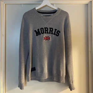Grå stickad tröja från Morris - Snygg grå stickad tröja från Morris med svart broderad logga och brittisk flagga framtill. Perfekt för dig som gillar klassisk och stilren look. I fint skick.