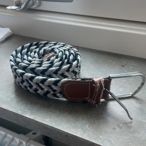 Randigt flätat bälte med läderdetaljer från Onewear! - Snyggt flätat bälte i svart och vitt med bruna läderdetaljer vid spännet och änden. Bältet har ett klassiskt silverfärgat metallspänne och passar perfekt till jeans. Den randiga designen ger en cool och modern look.