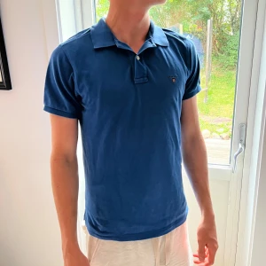 Blå pikétröja från Gant - Snygg blå pikétröja från Gant med klassisk krage och två knappar framtill. Tröjan har korta ärmar och Gant-logga broderad på bröstet. Perfekt för en chill och stilren look. Tillverkad i mjuk bomull som känns skön mot huden.