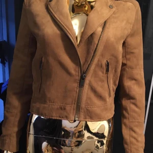 Brun tunn jacka H&M Divided strl 32 - Snygg tunn bikerjacka från H&M Divided i mocka imitation. Jackan har klassisk krage med tryckknappar, sned dragkedja och två fickor med dragkedja. Är i 100% Polyester Perfekt för dig som vill ha en cool och stilren look till vår och sommar 🙂