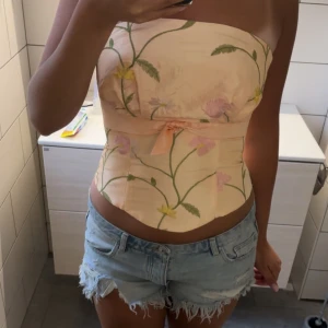 Blommig korsett med rosett - Superfin korsett i ljusbeige med broderade blommor i grönt, gult och rosa. Toppen är axelbandslös och har en söt liten rosett framtill. Perfekt för dig som vill sticka ut med en romantisk vibe.