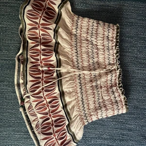 Beige mönstrad bandeau topp från Zara - Supersöt bandeau kjol från Zara i beige med röda och svarta broderade detaljer och elastisk smock. Kjolen har ett unikt bladmönster, knytband med pärlor och volangkant. Inbyggda shorts!