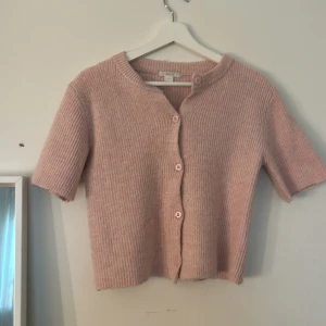 Rosa ribbad kofta med kort ärm - Superfin ljusrosa kofta med korta ärmar och ribbad stickning. Modellen är croppad och har knappar framtill. Perfekt att slänga över ett linne eller t-shirt för en söt och chill look.